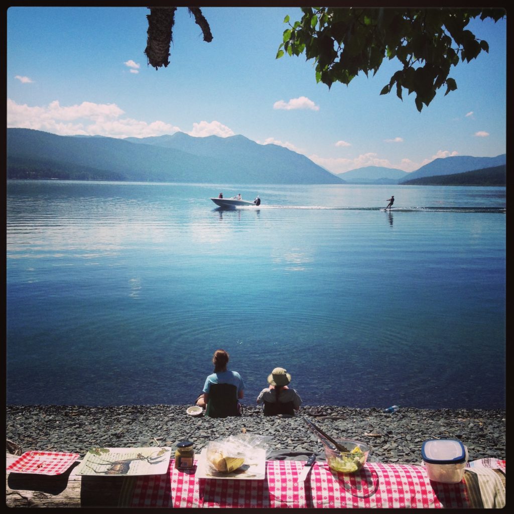 A checkered tablecloth on a log onthe shores of a lake with two people watching watching a paddleboarder glide by 