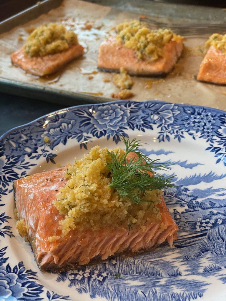 Parchment paper lined rimmed baking sheet with 4 pieces of herb coated steelhead trout next to a blue plate of bake trout