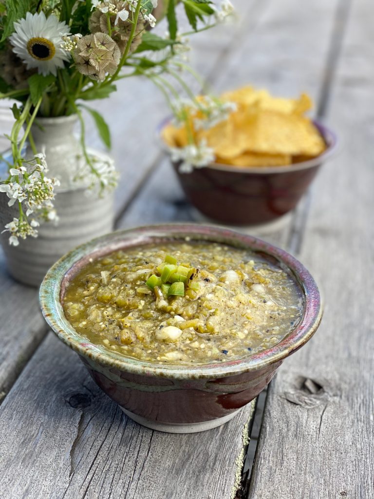 a colorful green and purple ceramic bowl full of salsa verde next to a similar bowl full of tortilla chips and a white vase of flowers