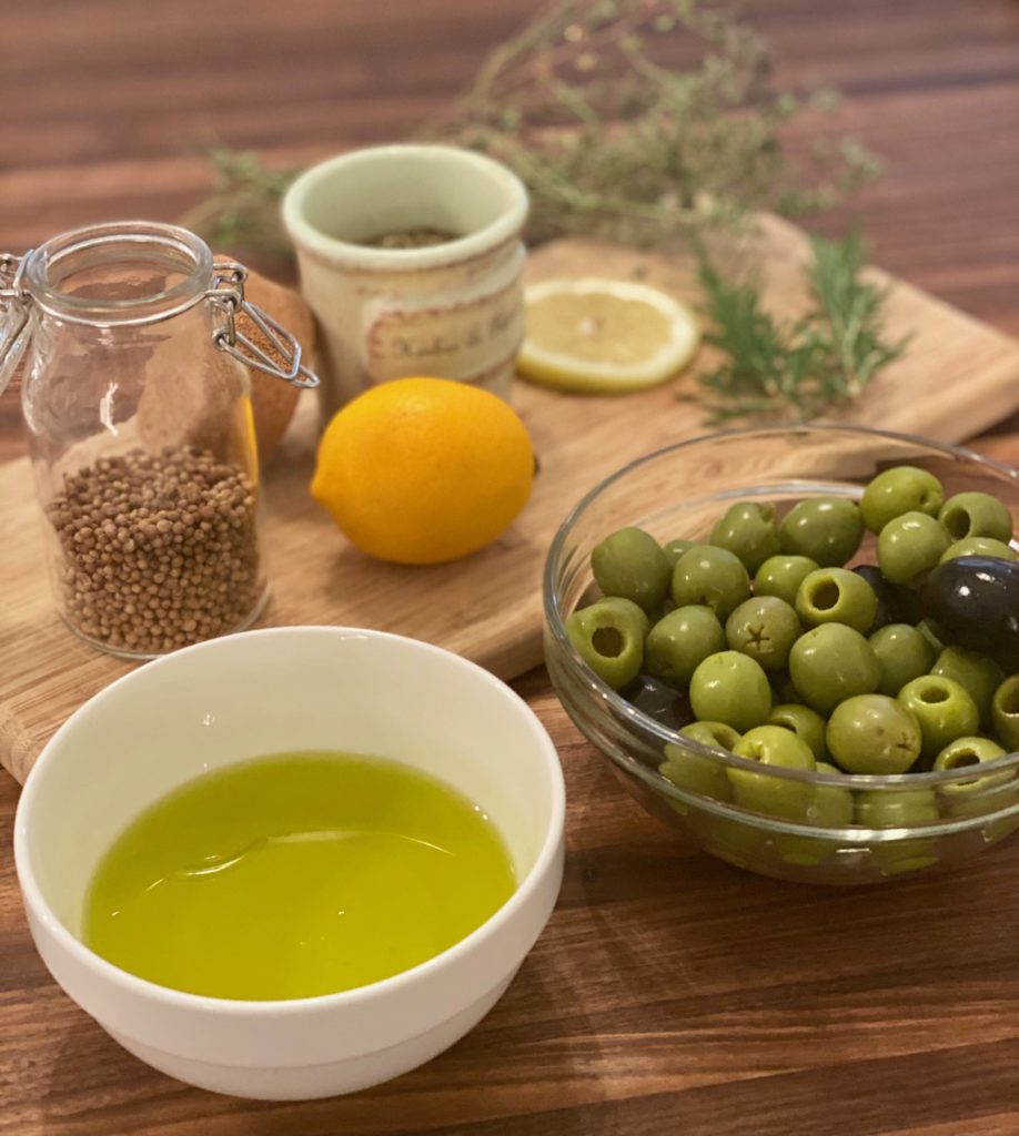 a wooden tray of coriander seeds in a glass jar, a ceramic jar of herbs, a lemon and a glass bowl of green olives and a bowl of olive oil