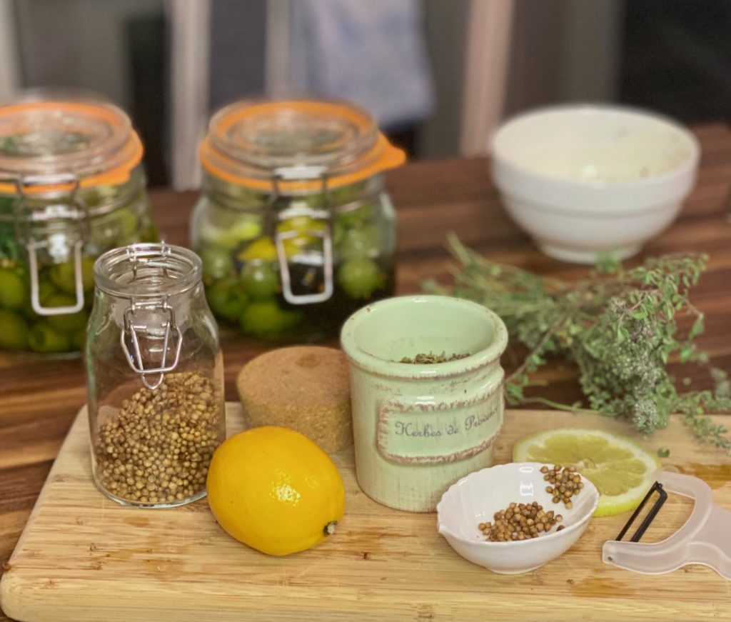 a wooden tray of coriander seeds in a glass jar, a ceramic jar of herbs, a lemon and two jars of green olives 