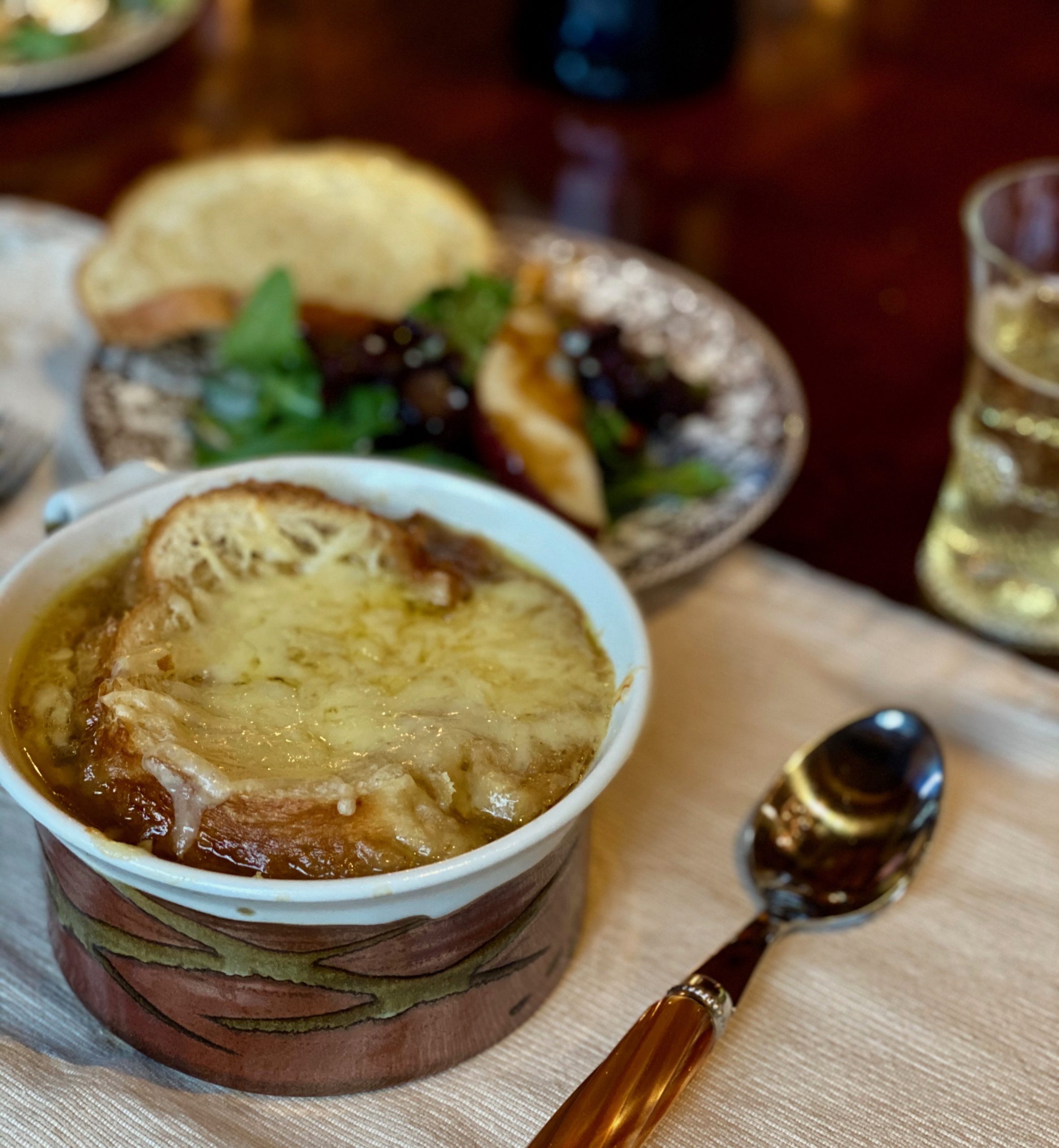 A justice brown ceramic soup bowl with handle filled with cheese topped soup next to a spoon and a plate of green salad and toast
