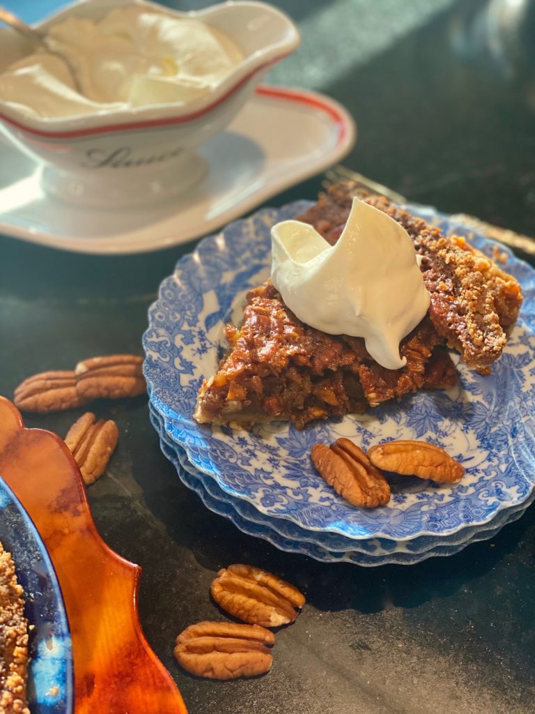Bourbon pecan pie with a dollop of whipped cream on top, on a stack of 3 blue china plates next to some pecans and a dish of whipped cream