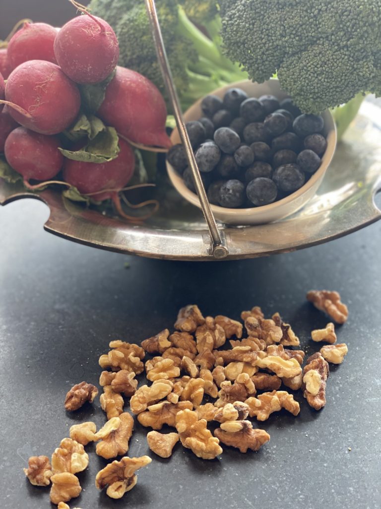 A silver dish with blueberries, rashes and a head of broccoli next to  a pile of walnuts