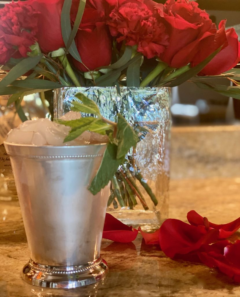 a frosted silver julep cup filled with ice next to a vase of red roses 