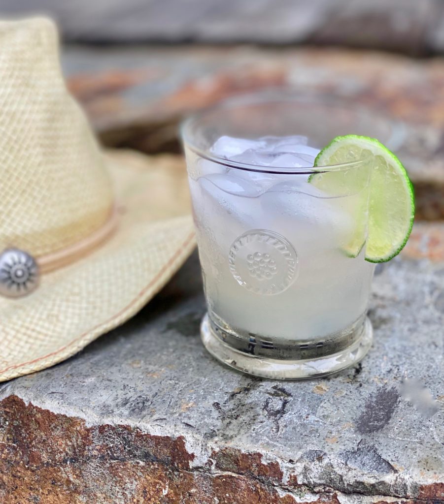 a clear glass filled with a frosty cold drink garnished with a lime wedge sitting on a flat stone next to a straw cowgirl hat 
