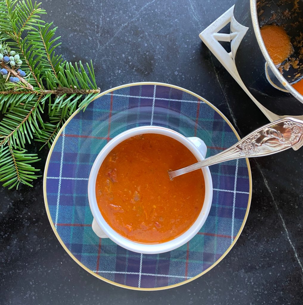 a blue tartan plate with a white rimmed bowl filled with tomato soup with a silver spoon insisted in the soup