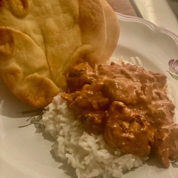 A white plate with a red tomato sauce, chicken over a bed of cooked rice and slices of naan bread