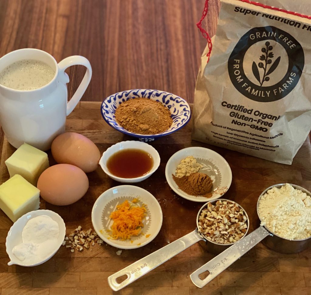 ingredients for pancakes on a wooden bowl- a brown bag of flour, eggs, spices on small dishes, two chunks of butter, a white small pitcher of milk, and measuring cups filled with chopped nuts and flour