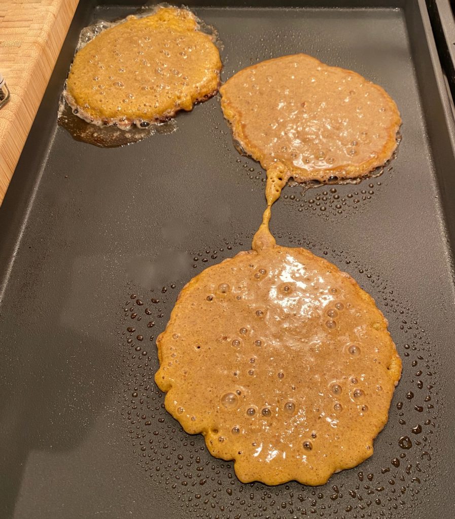 A griddle with three buckwheat pancakes cooking 