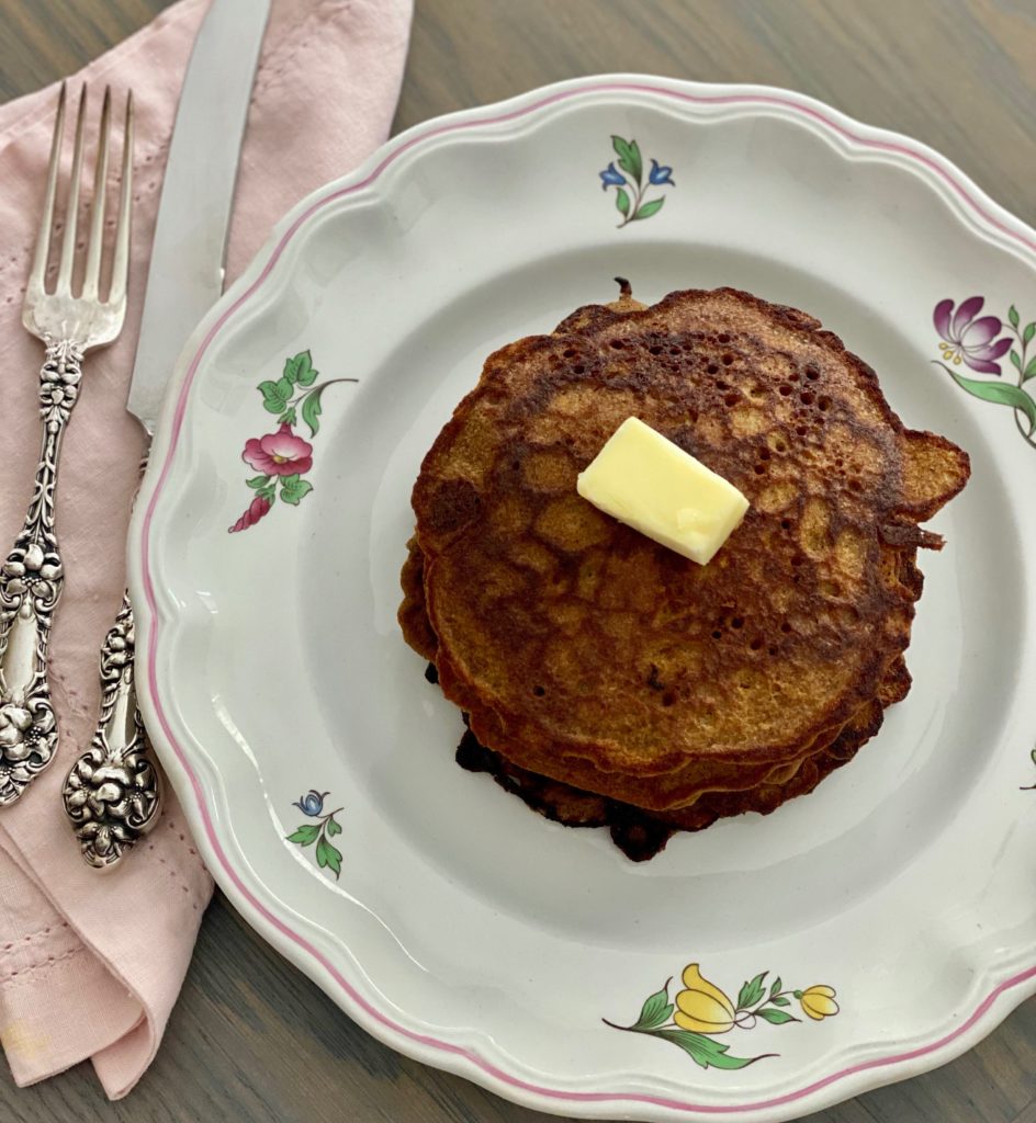 A white plate with flowers with a stack of pancakes, a pat of butter and an ornate sterling silver fork and knife resting on a pink napkin off to the left side.