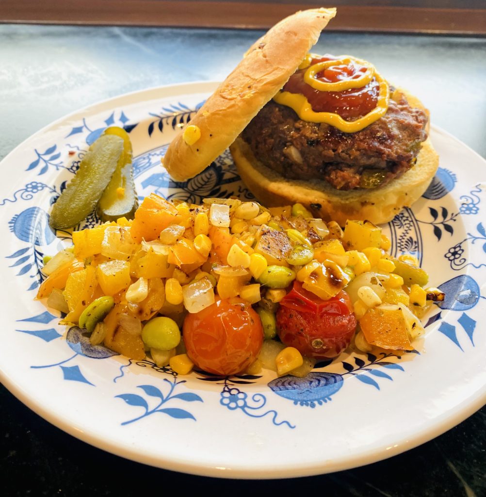 a burger on a blue and white plate with a pile of fresh corn and tomatoes
