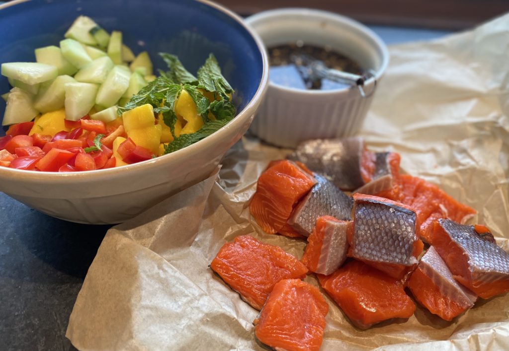 parchment paper with raw salmon chunks, a bowl of chopped vegetables and a soy sauce 