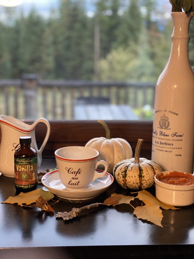 a white bistro coffee cup and saucer, a tall porcelain jug and bottle of vanilla two small white pumpkins and a small bowl of pumpkin puree staged in front of a window