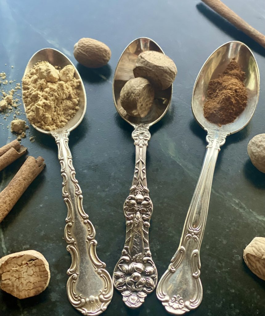 Three antique silver teaspoons filled with powdered ginger, whole nutmeg and ground cinnamon surrounded by other whole spices on a counter. 