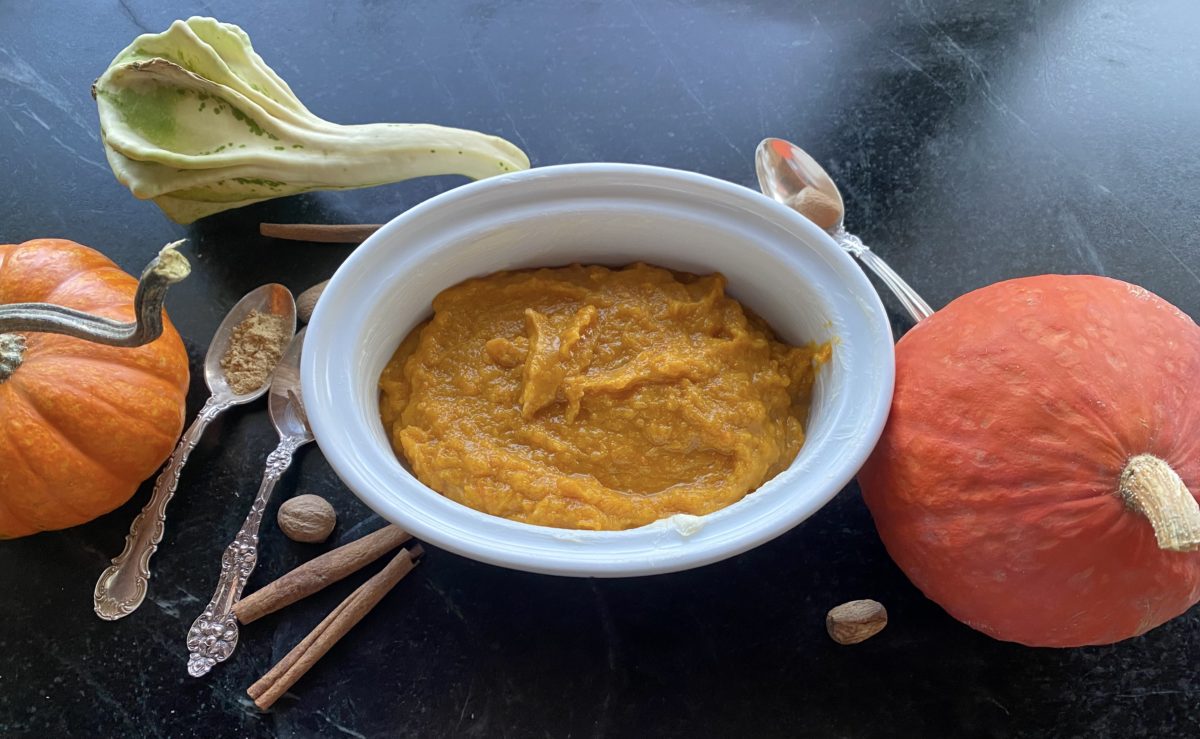 A baked squash casserole in a dish surrounded by silver spoons, whole spices and a variety of autumn gourds