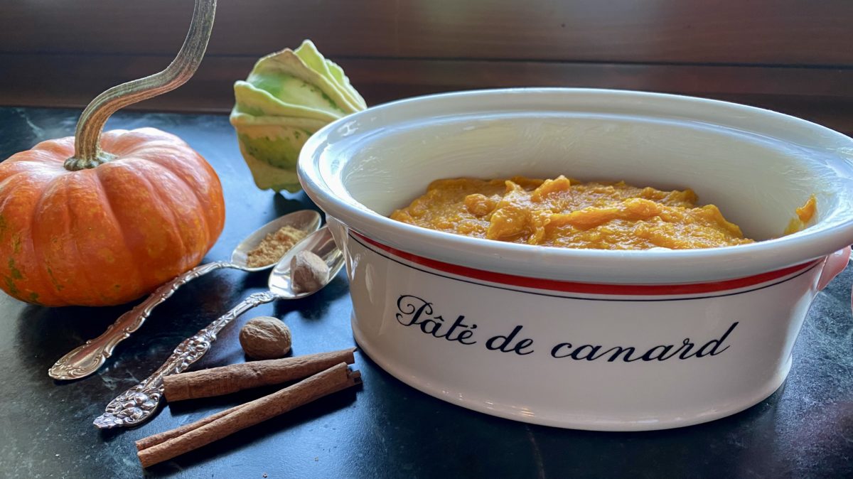 A deep casserole dish filled with mashed orange butternut squash surrounded by a stemmed pumpkin and spoons and whole nutmeg and cinnamon sticks