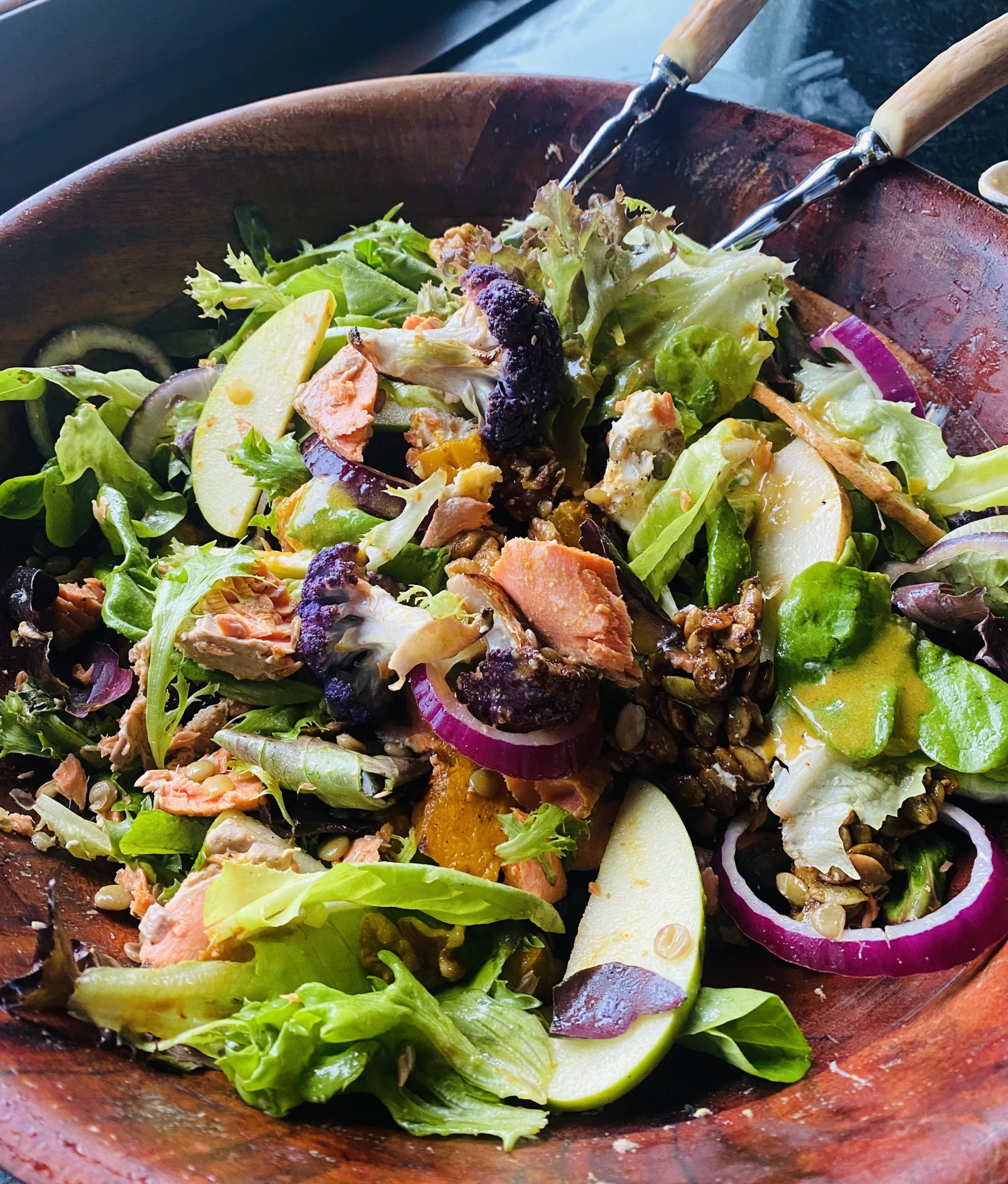 a wooden bowl with salad tongs surrounded by a green salad filled with pears, salmon and onions