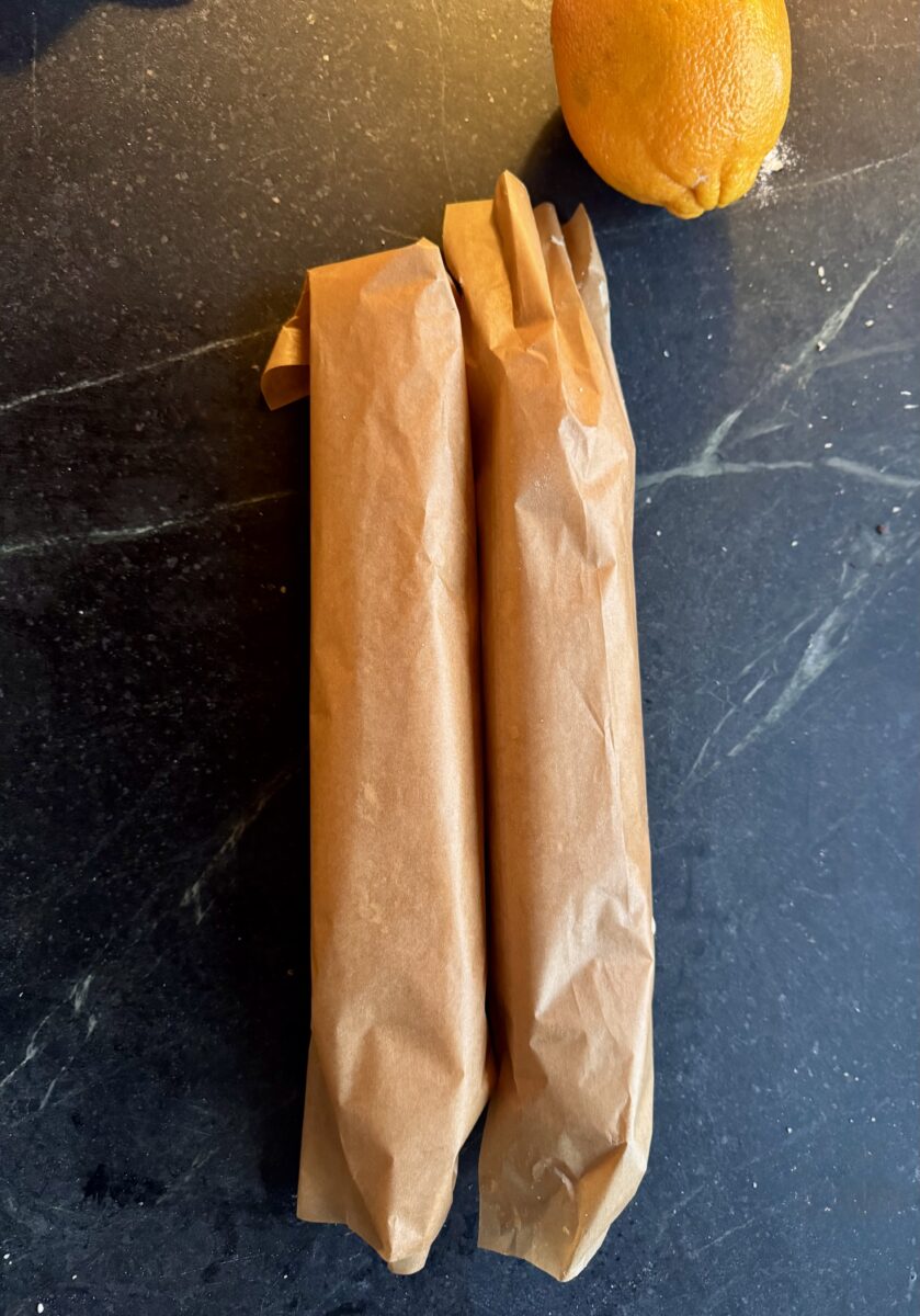 two long rolls of shortbread wrapped tightly in brown parchment paper on a soapstone counter next to an orange 