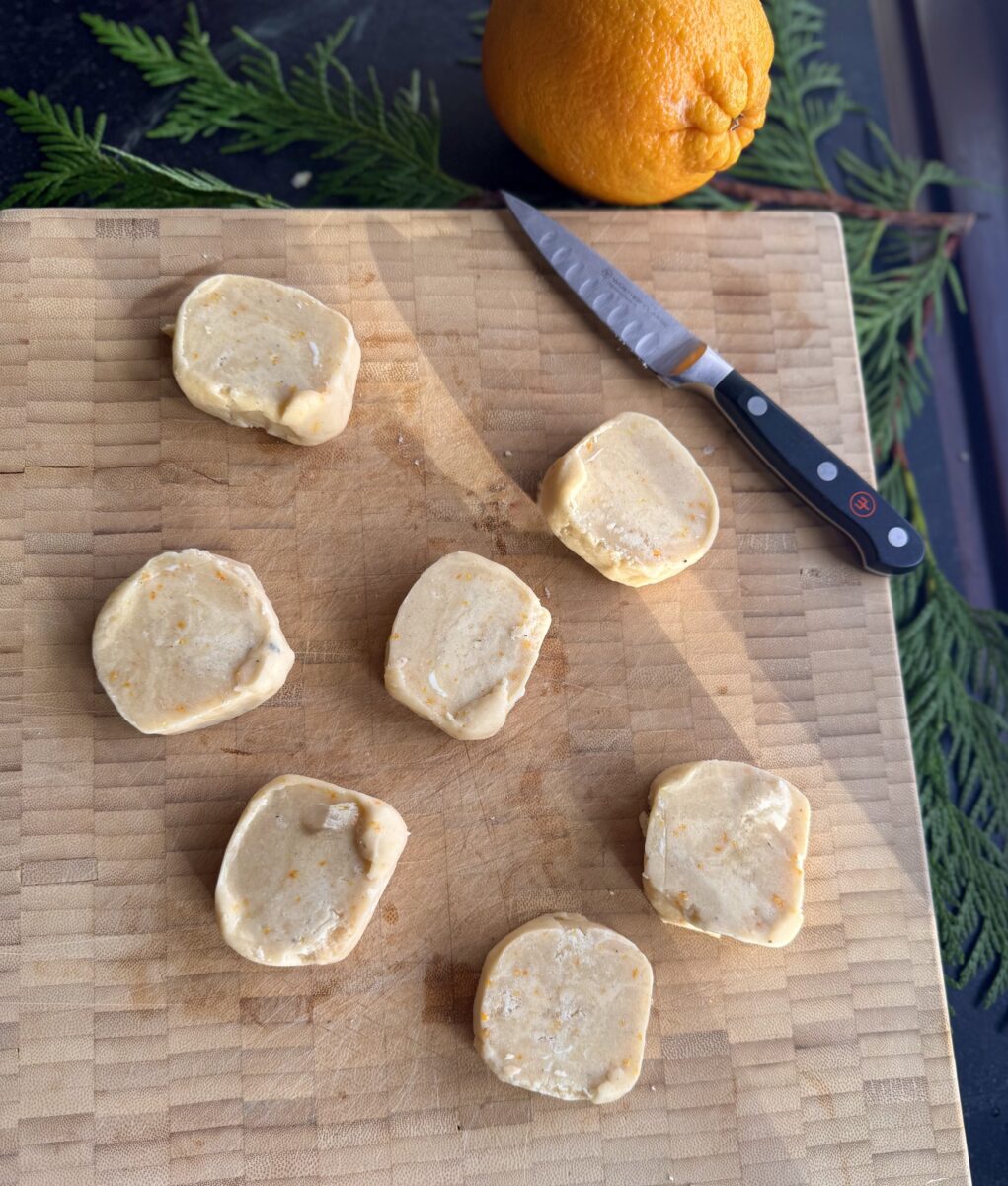 tray of unbaked orange shortbread cookie slices on a cutting board next to a small pairing knife and a whole orange 
