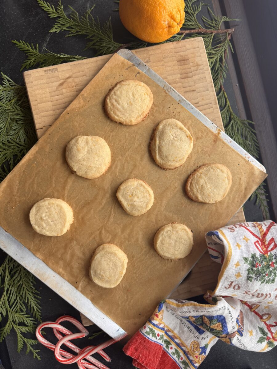 a cookie sheet lined with brown parchment paper holding 8 lightly browned baked orange shortbread cookies next to a holiday dishtowel and a whole orange and peppermint candy canes
