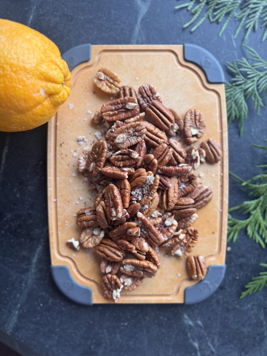 halved pecans on a cutting board next to pieces of cedar evergreens and a whole orange 