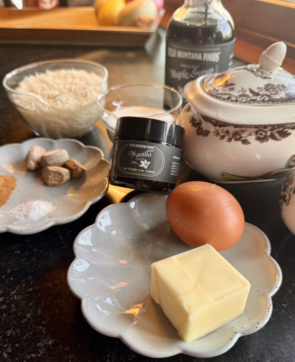 ingredients for vanilla rice pudding - an egg on a grey plate, sugar, a jar of vanilla paste, a cup of rice in a clear glass bowl and a pile of whole nutmeg 