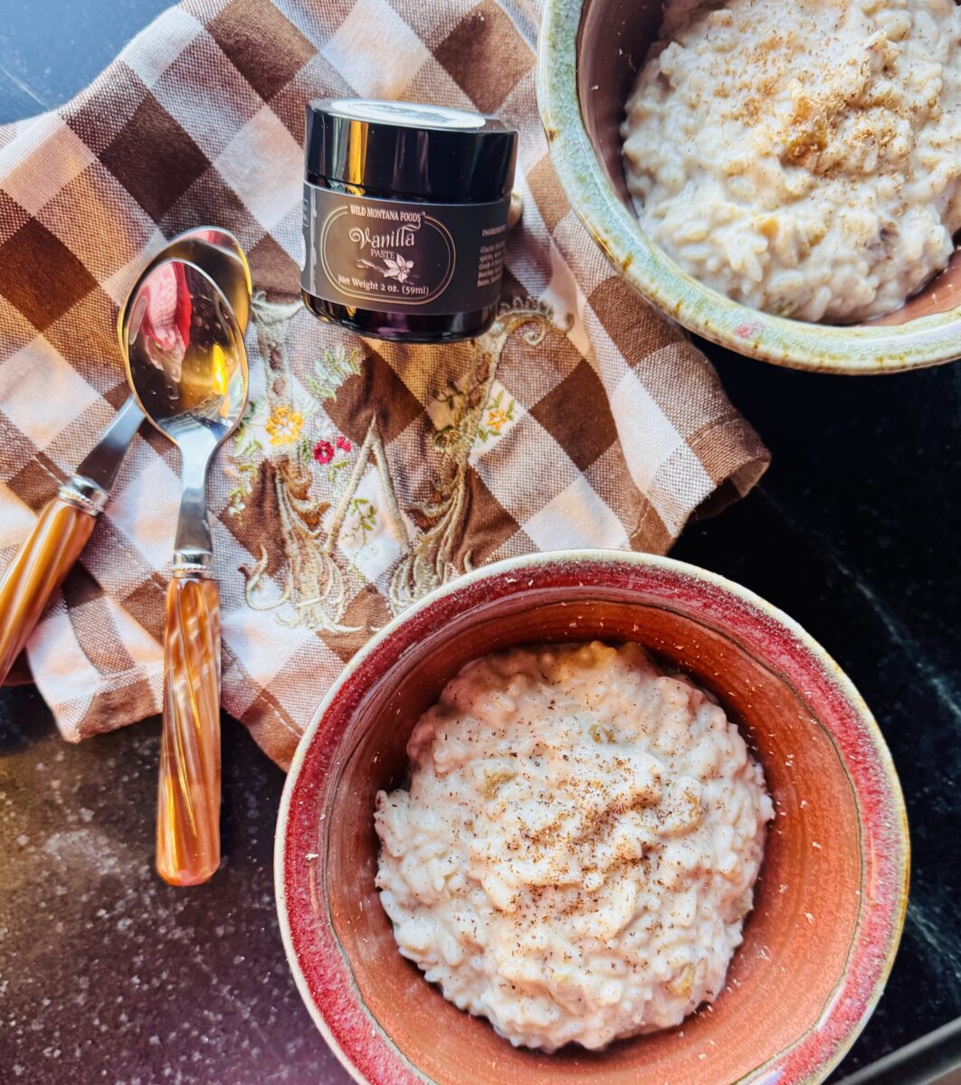 Two earthenware bowls filled with vanilla bean rice pudding, two spoons, a jar of vanilla bean paste all resting on a brown and white buffalo checked napkin