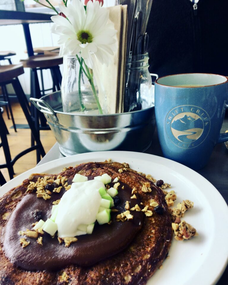 a pile of pumpkin pancakes on a white plate next to an earthenware mug of coffee and a vase of daisies