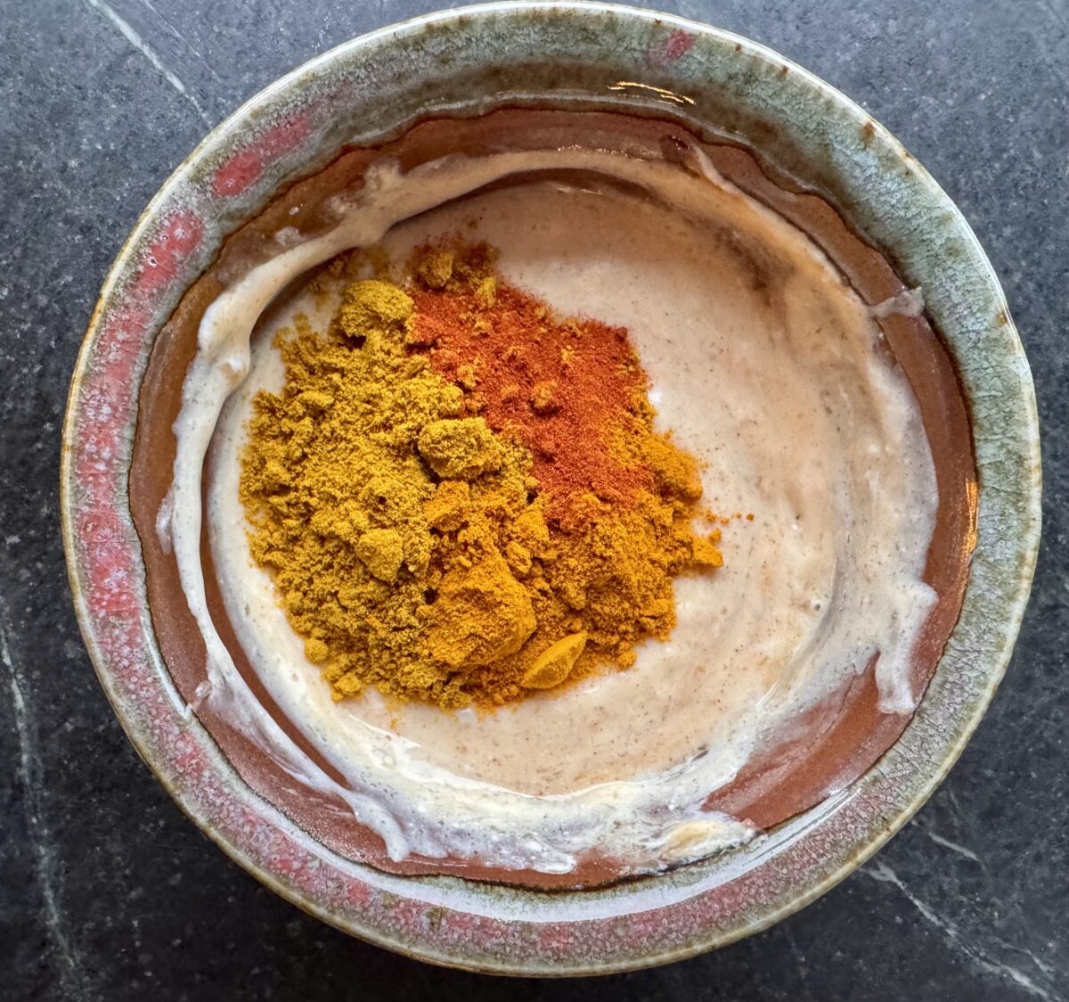 a bowl of mayonnaise and yogurt in an earthenware bowl with a mound of curry and turmeric on top of the mixture for a curry chicken salad recipe 