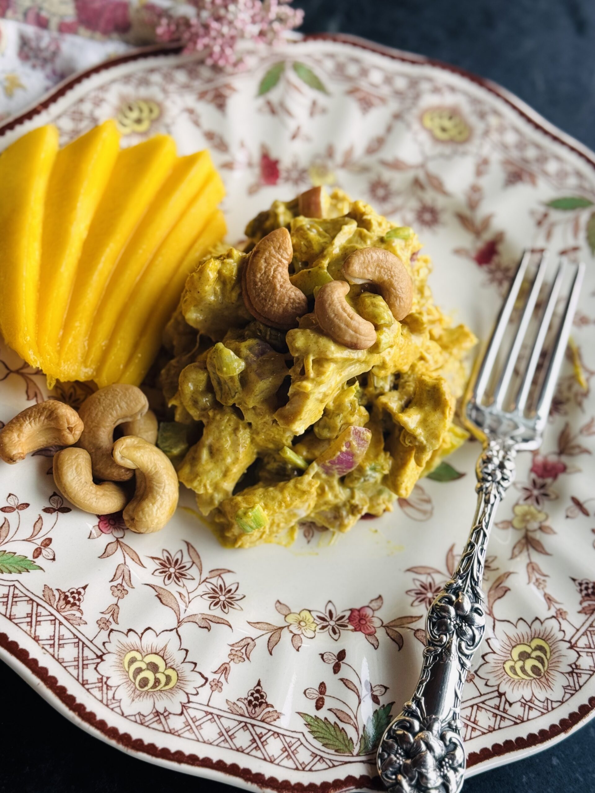 An antique brown transfer ware plate with a scoop of curry chicken salad on it, sliced mangos and an antique sterling silver fork