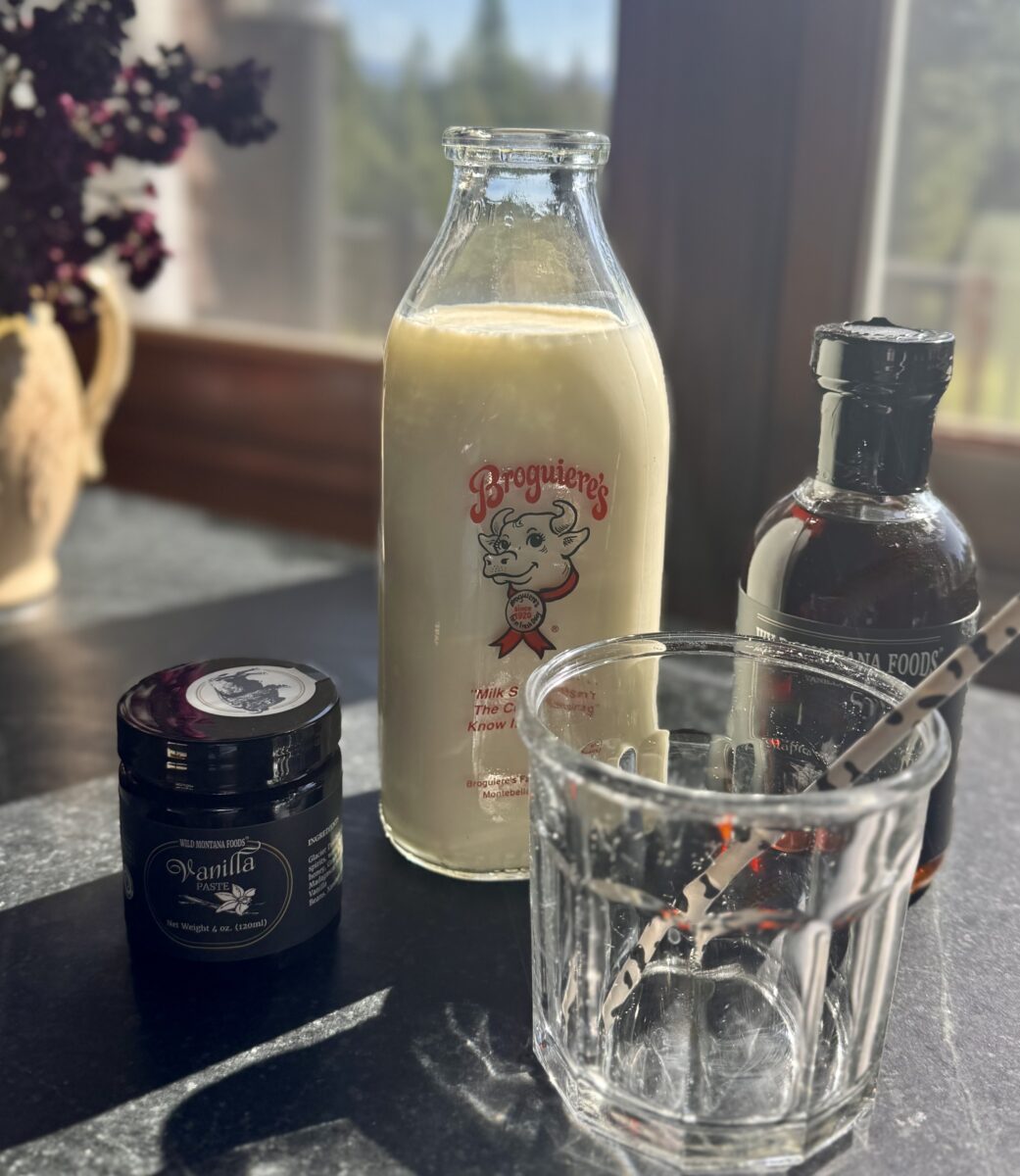 a glass next to a jar of vanilla bean paste and a bottle of vanilla maple syrup. In the background is a jug of milk