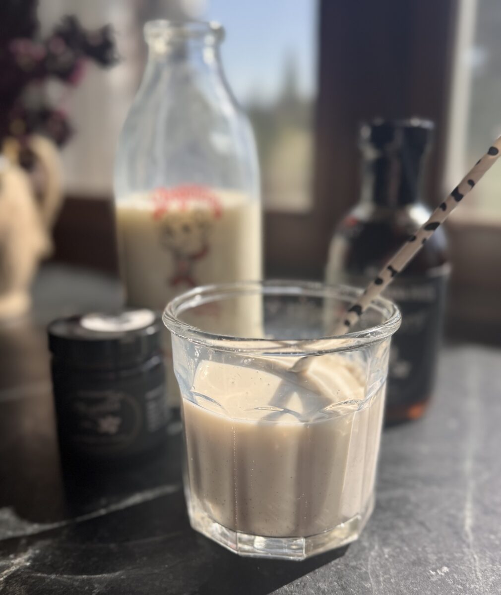 a small glass of vanilla bean milk with a straw and a jar of vanilla bean paste and a bottle of vanilla bean maple syrup and in the background is an antique glass milk bottle filled with milk
