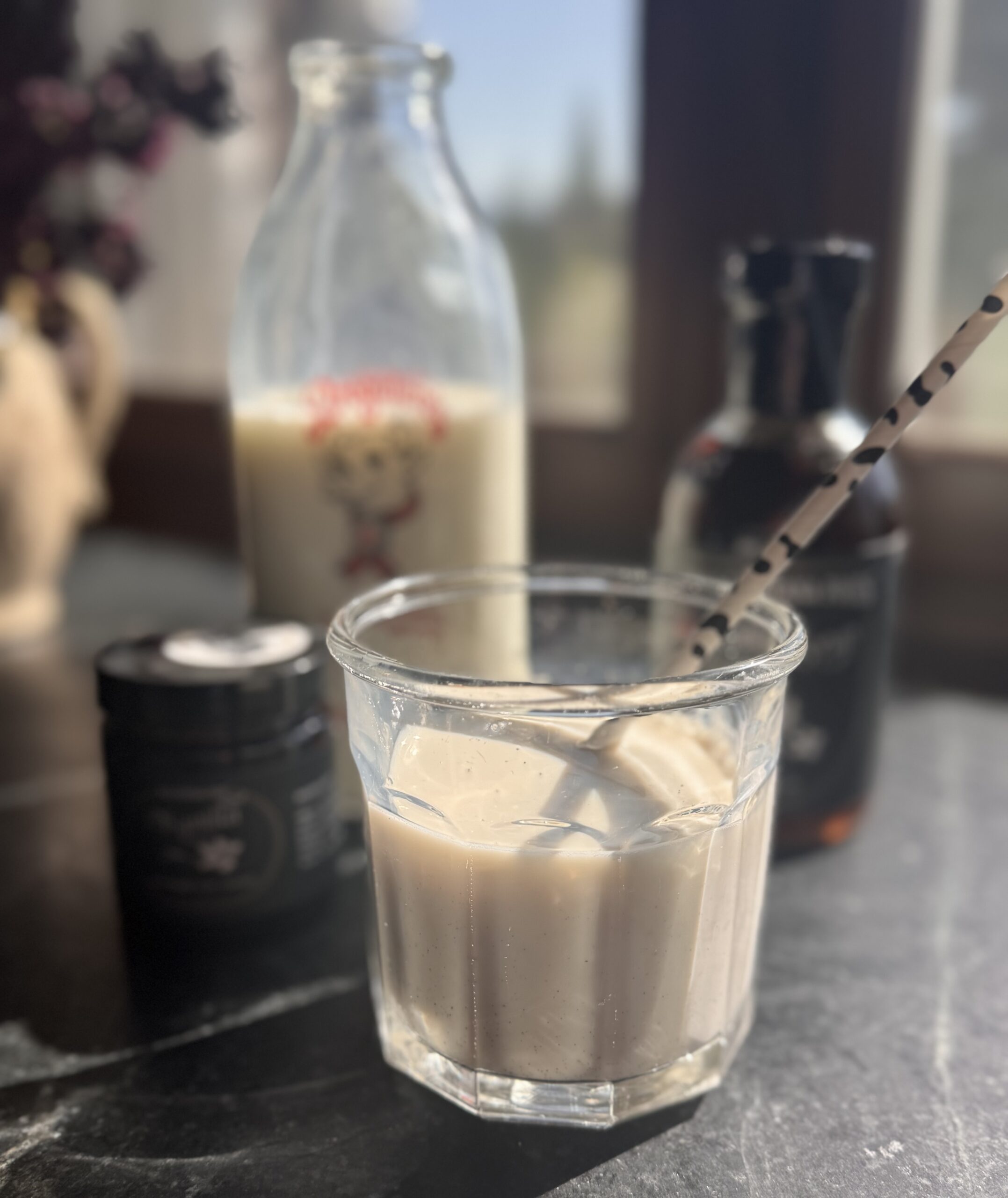 a small glass of vanilla bean milk with a straw and a jar of vanilla bean paste and a bottle of vanilla bean maple syrup and in the background is an antique glass milk bottle filled with milk